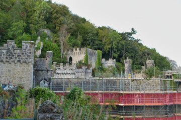 A weathered stone castle ruin with turrets and green foliage sits beneath a wooded hill. Scaffolding is visible in the foreground - Gwrych Castle, Abergele, Wales, UK © Robert