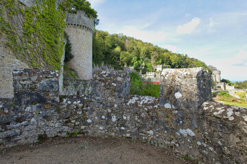 A close-up view of a weathered stone castle wall with battlements and a round tower, showing a distant forested hill under a blue sky - Gwrych Castle, Abergele, Wales, UK © Robert