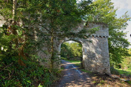 A weathered stone archway, part of a castle ruin, spans a narrow path through lush greenery on a sunny day - Gwrych Castle, Abergele, Wales, UK