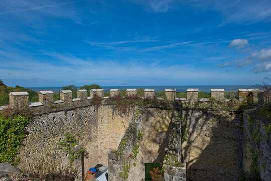 A sunlit view from ancient castle ruins shows crenelated stone walls, overgrown with plants, extending towards a bright blue sea under a clear sky - Gwrych Castle, Abergele, Wales, UK