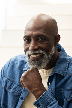 Senior African man resting chin on fist, smiling on pale staircase in denim jacket, showing ring