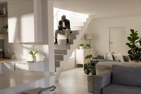 Senior African American man sitting on floating stairs holding mug, looking toward white cabinets