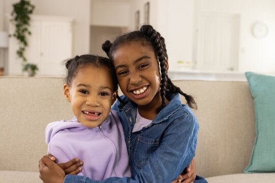 African American child sisters hugging on beige sofa wearing purple hoodie and denim jacket