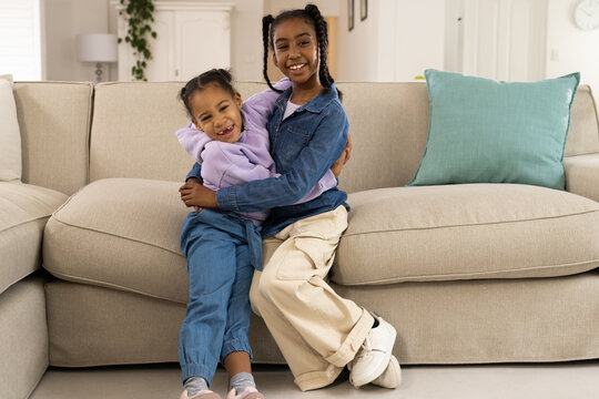 African American female youths sitting on beige sectional sofa hugging and smiling in living room