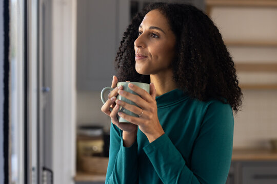 African American woman standing in kitchen by window holding ceramic mug, wearing teal top and ring
