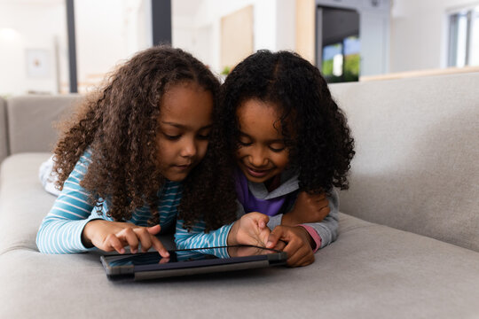 African American female youth friends lying side-by-side on light-gray sofa, using black tablet