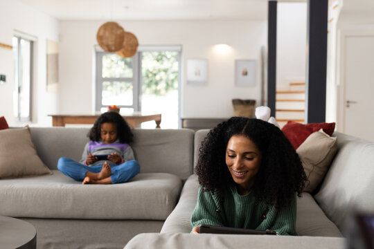 African American mother and daughter relaxing on sofas in living room using tablet handheld device