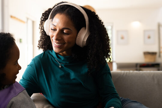 African American mother and daughter sitting on living room couch, wearing white headphones