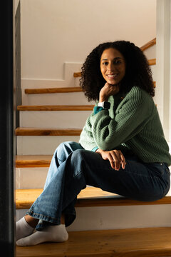 African American woman sitting on wooden steps at home in sweater checking smartwatch near handrail