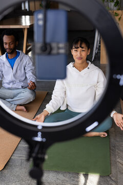 African American couple in hoodies sitting on green mat at home recording with ring light phone