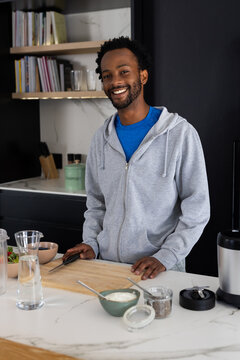 Man in his thirties wearing gray hoodie blue tee in home kitchen holding knife on board