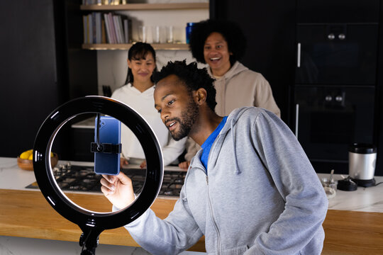 Diverse friends adjusting smartphone on ring light at wooden kitchen island wearing hoodies