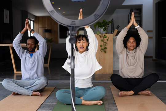 Diverse friends practicing yoga on mats, sitting cross-legged at home with ring light, camera