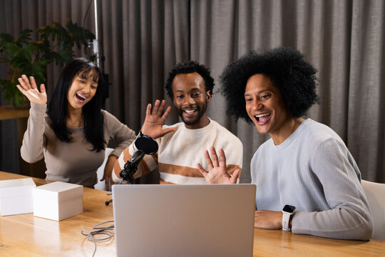 Diverse coworkers smiling and waving at laptop in home studio with microphone and white boxes