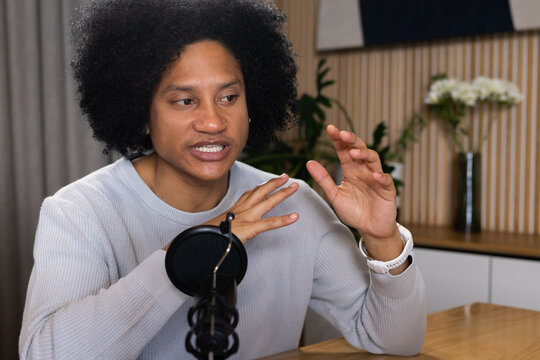 Adult male speaker wearing gray sweater speaking into mic and gesturing at studio table