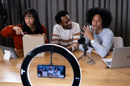 Co-hosts wearing knit sweaters opening cardboard box with scissors at wooden table under ring light