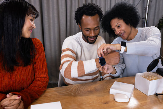 Diverse friends sitting at wooden table in curtained room testing smartwatches, boxes, mic arm