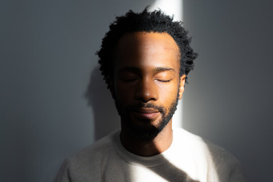 African man sitting eyes closed, bathing in narrow sunbeam, shadow falling on gray wall, sweater