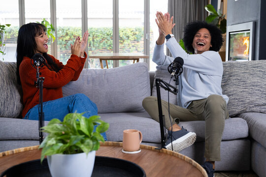 African American co-hosts laughing and clapping while recording on sofa in home with microphones