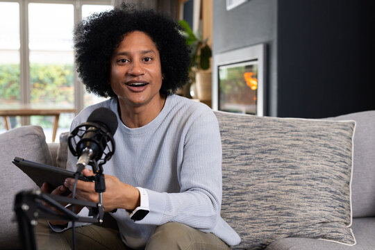 African American man sitting on gray sofa in home studio speaking into microphone holding tablet