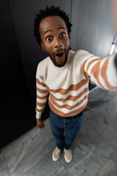 African American man standing in narrow modern hallway holding smartphone and wearing cream sweater