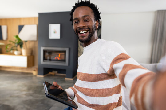 African American man sitting on light-colored sofa near modern fireplace holding tablet smiling