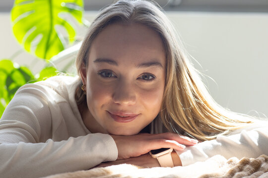 Woman lying on plush blanket, resting chin on hands, smiling by window with Monstera and smartwatch