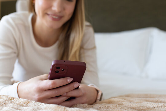 Adult woman reclining on bed, holding burgundy smartphone while wearing smartwatch and light top