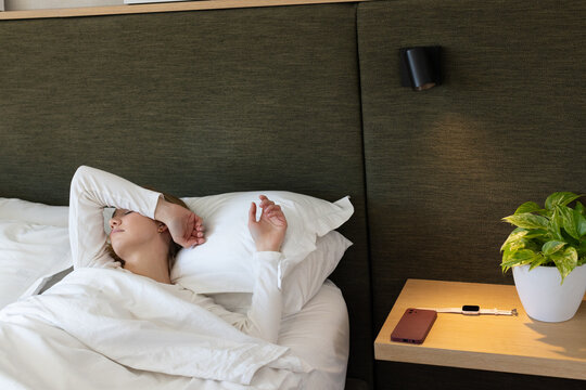 Adult female lying in bed, resting with duvet, headboard, table with phone and charging smartwatch