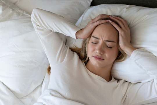 Woman lying on bed pressing both hands to temples, wearing white top among pillows, copy space