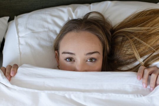 Adult female lying in bed with white duvet pulled up, holding sheet and showing gold earring