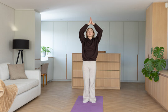 Female pressing palms together overhead, standing on purple mat in living room in sweats