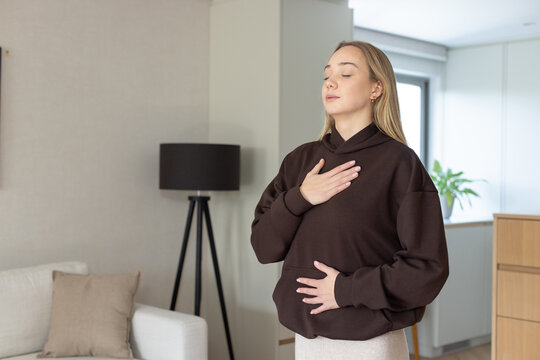Woman holding hands on chest and belly, wearing hoodie, standing in living room near lamp, plant