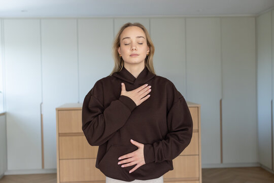 Woman standing in bedroom wearing hoodie and pants, hands on chest, dresser and wardrobe behind