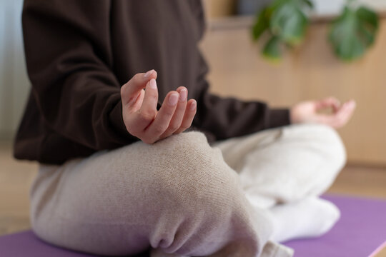 Female in brown top, beige sweatpants meditating on purple yoga mat at home near wooden cabinet