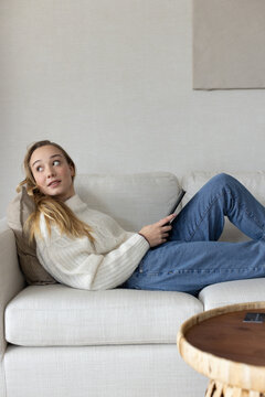 Woman reclining on cream sofa wearing cable-knit sweater, jeans holding black tablet, coffee table