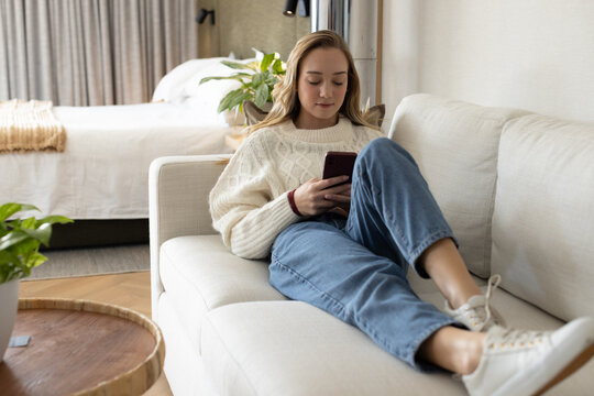 Woman reclining on light sofa at home, scrolling smartphone while wearing cream sweater and jeans