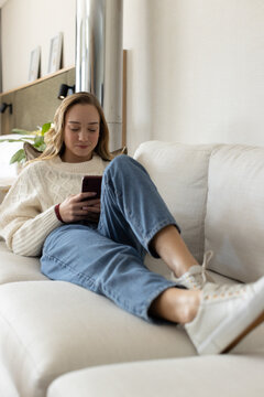Adult female reclining on light beige sofa at home, holding smartphone and wearing cream sweater