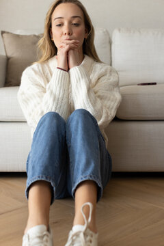 Adult female sitting on herringbone floor hugging knees in cream sweater leaning by beige sofa