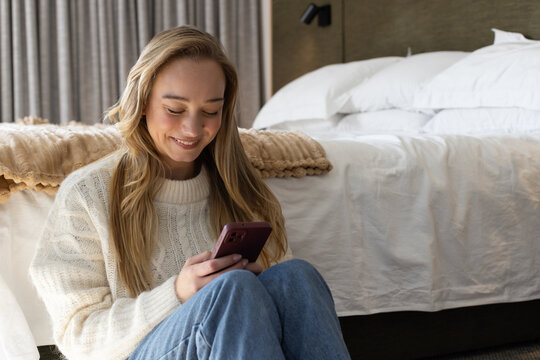 Female in cream sweater and jeans sitting on floor in bedroom holding mauve smartphone, smiling