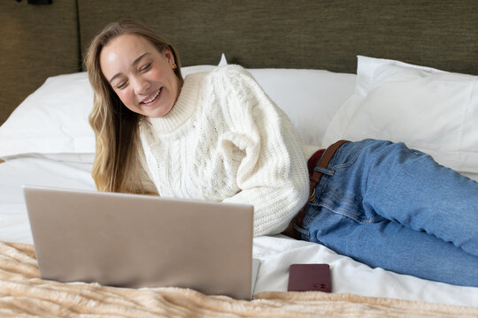 Adult woman reclining on bed using laptop and smartphone, wearing cable knit sweater and jeans