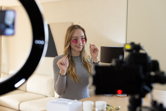 Female host in gray top wearing pink undereye patches filming skincare ring light camera at home
