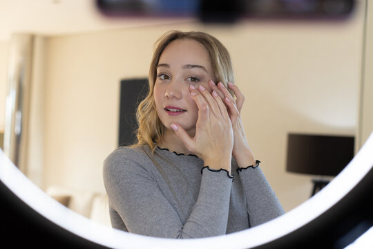 Woman wearing grey top touching below right eye checking mirror at home with ring light