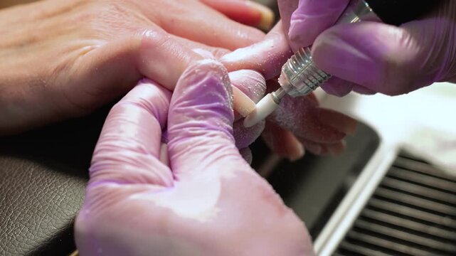 A horizontal slow motion macro video showing a manicurist using an electric drill cutter to carefully shape a client's fingernail in a beauty salon.