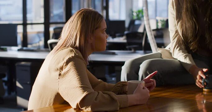 Diverse female coworkers sitting at office table, second woman placing mug, talking about work