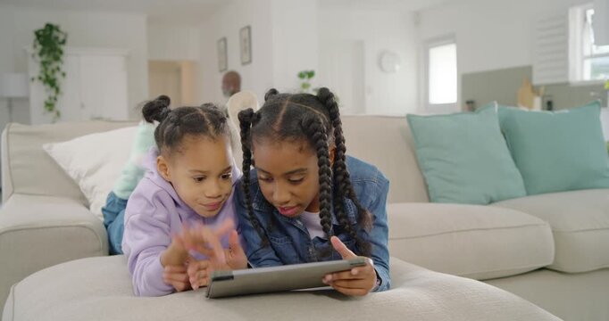 Two youth girls lying on ottoman in living room sharing tablet watching one pointing other reacting