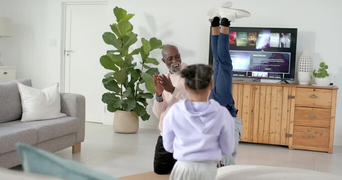 African man with two kids clapping, cheering, urging child kicking headstand in living room by TV