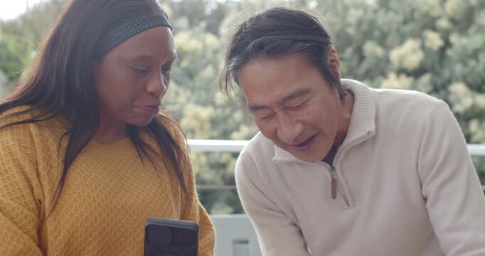 Senior Asian man and African woman showing smartphone design and marking pumpkins on balcony