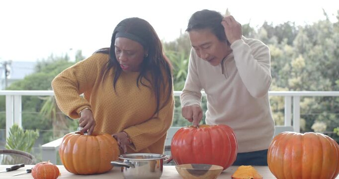Cutting pumpkins, Senior Asian man, African woman scooping pulp into metal pot for soup on balcony