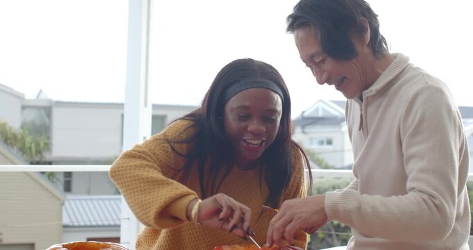 Senior Asian man, African woman scooping pumpkin pulp on balcony after removing tops, filling pot